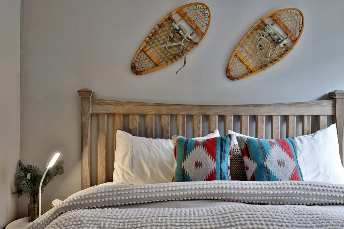 A tranquil bedroom scene featuring soft bedding, colorful patterned pillows, and snowshoes hung on the wall. The space radiates comfort and invites relaxation, with natural light filtering in to enhance the serene ambiance.