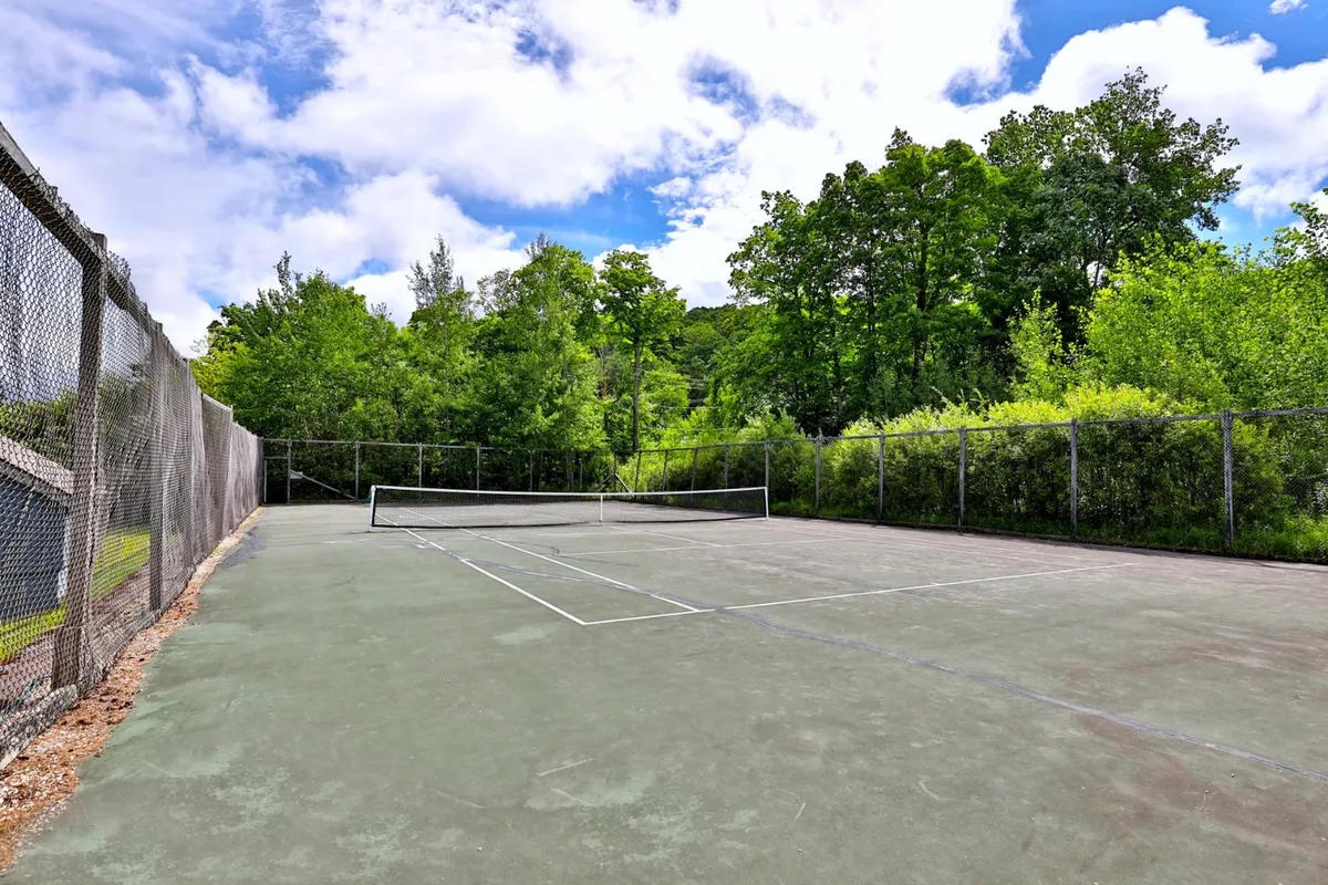 This image depicts an exterior tennis court surrounded by lush greenery. The net is taut and ready for play, with trees and the natural landscape of Vermont visible in the background, inviting outdoor activity and enjoyment.