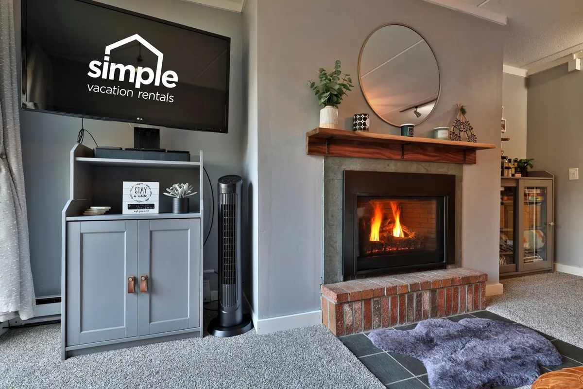 A cozy living space anchored by a crackling fireplace, with a television above the mantel, decorative plants, and comfortable seating. The ambiance is warm and welcoming, ideal for family gatherings, movie nights, and relaxing after a day of exploration.