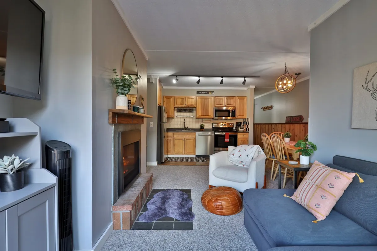 The image focuses on a homely living area with a stone fireplace, a plush sofa, a dining table, and an open view of the kitchen. The ambiance is warm and inviting, suggesting a perfect spot for cozy evenings, shared meals, and storytelling by the fire.