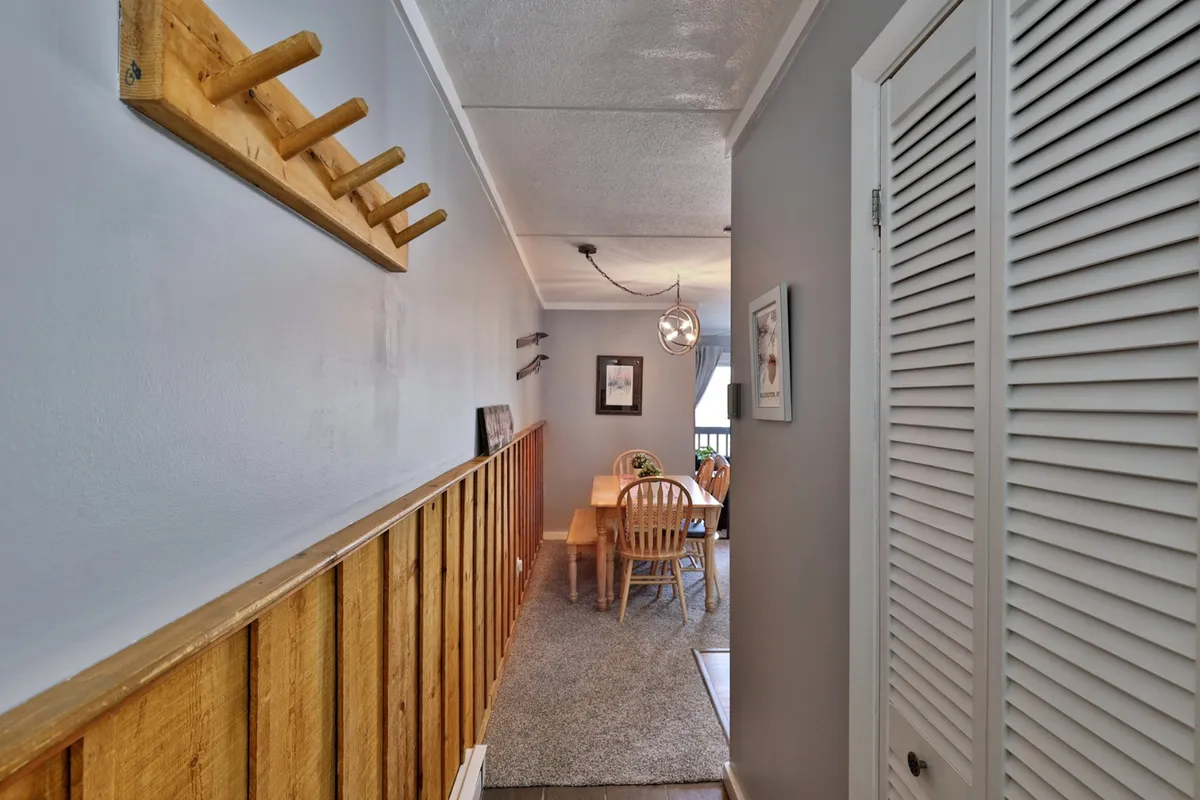 A cozy interior scene showing a dining table set near a wooden coat rack and illuminated by a warm light fixture. The window allows for natural light, hinting at views outside and evoking the idea of enjoying morning coffee while appreciating the tranquil woodland setting.