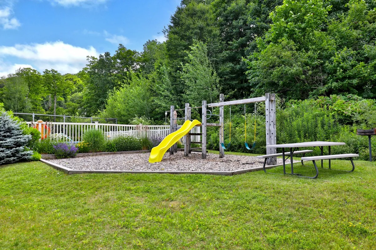 A cheerful outdoor play area featuring a yellow slide, swing set, and picnic table, all surrounded by lush greenery. The space is designed for children to play, families to bond, and to create memorable moments outdoors.