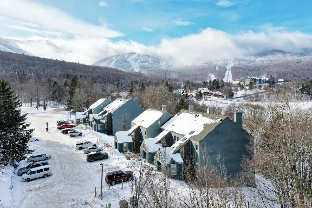 A wintry exterior scene with snow-covered roofs, a backdrop of majestic mountains, and vehicles parked in front of the cozy condo buildings. The scene evokes a sense of mountain adventure, comfort, and winter sports activities.