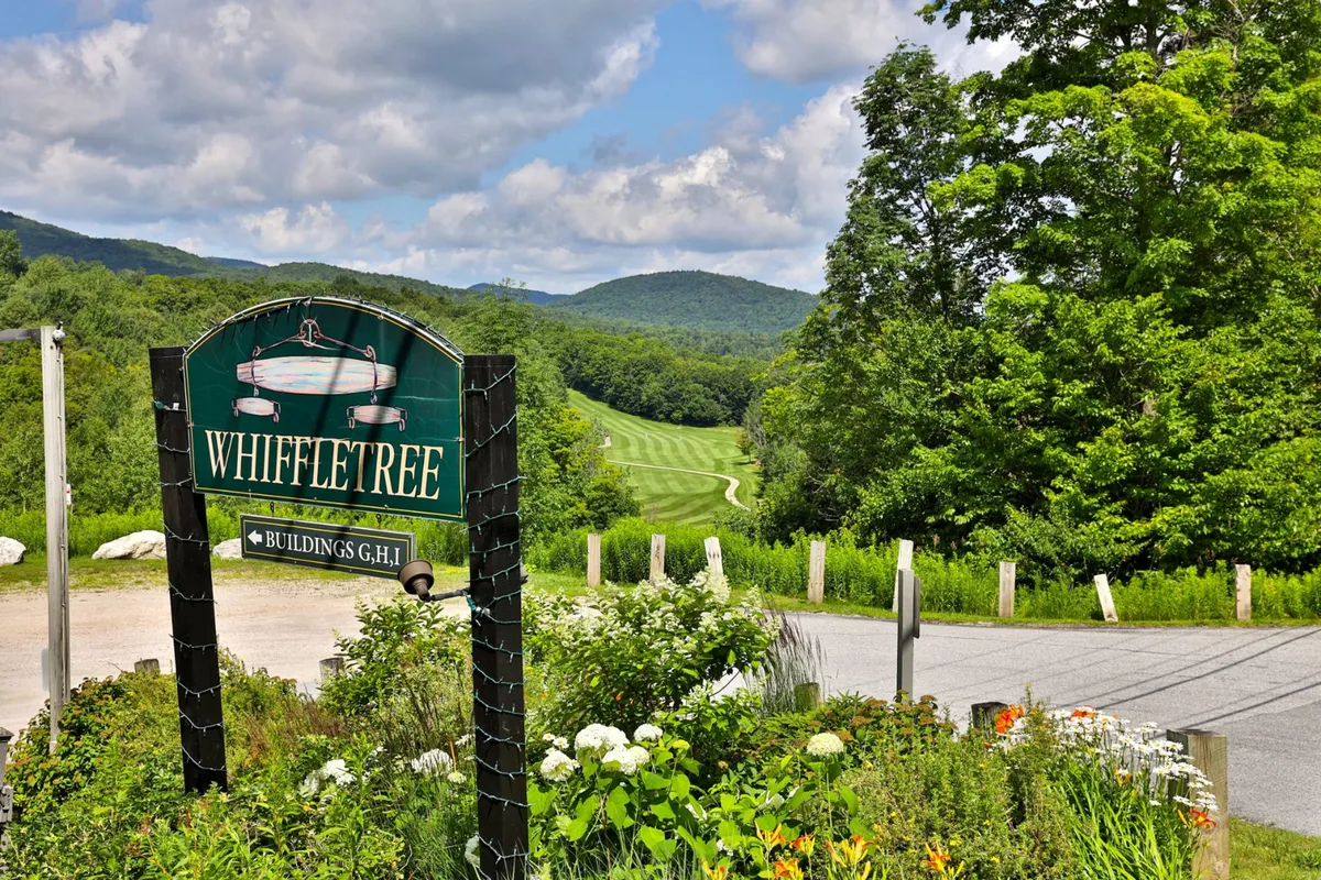 A welcoming sign for the Whiffletree property sits amid lush greenery, backed by a striking mountain landscape. The scene feels peaceful and inviting, suggesting easy access to the outdoors and a sense of arrival at a mountain retreat.