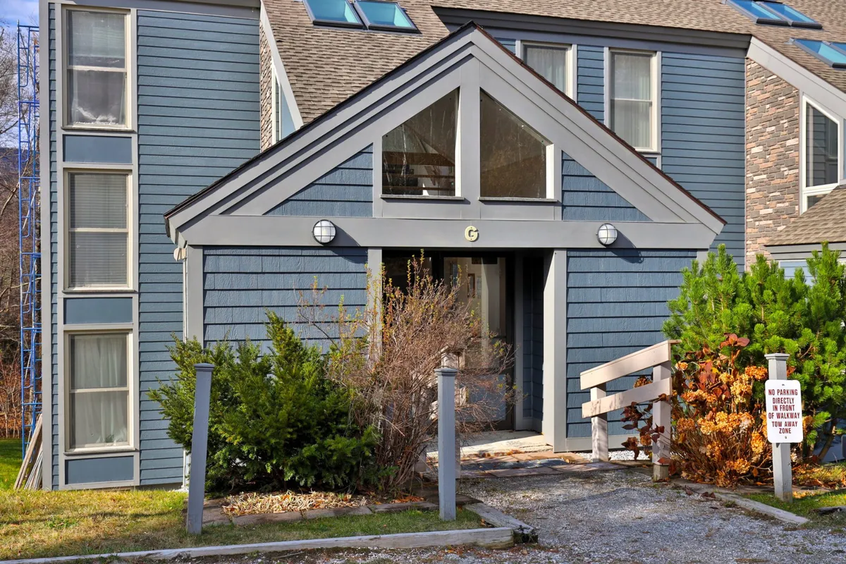 This exterior shot highlights the inviting triangular entrance canopy, textured blue siding, and surrounding landscaping. The welcoming front porch is subtly framed by snowfall, emphasizing the comfortable arrival experience during winter.