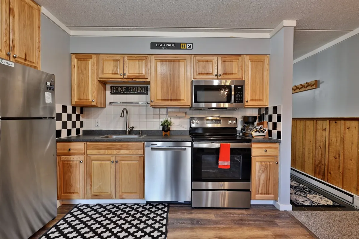 A warmly lit kitchen featuring stainless steel appliances, wood cabinetry, and a striking black and white tile backsplash. A decorative sign adds a touch of personality, while counters are well-stocked and spacious—ideal for meal prep or social cooking.